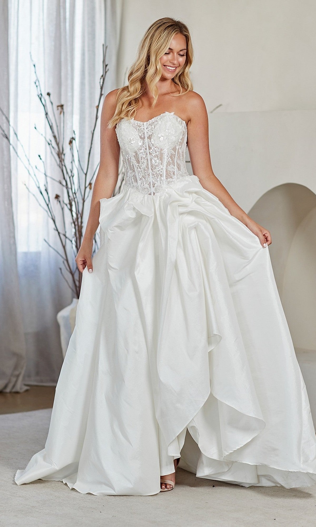 A woman stands indoors, smiling as she holds out the sides of her white Amelia Couture 2087 long prom dress, featuring a strapless lace corset bodice and a full, draped A-line skirt.